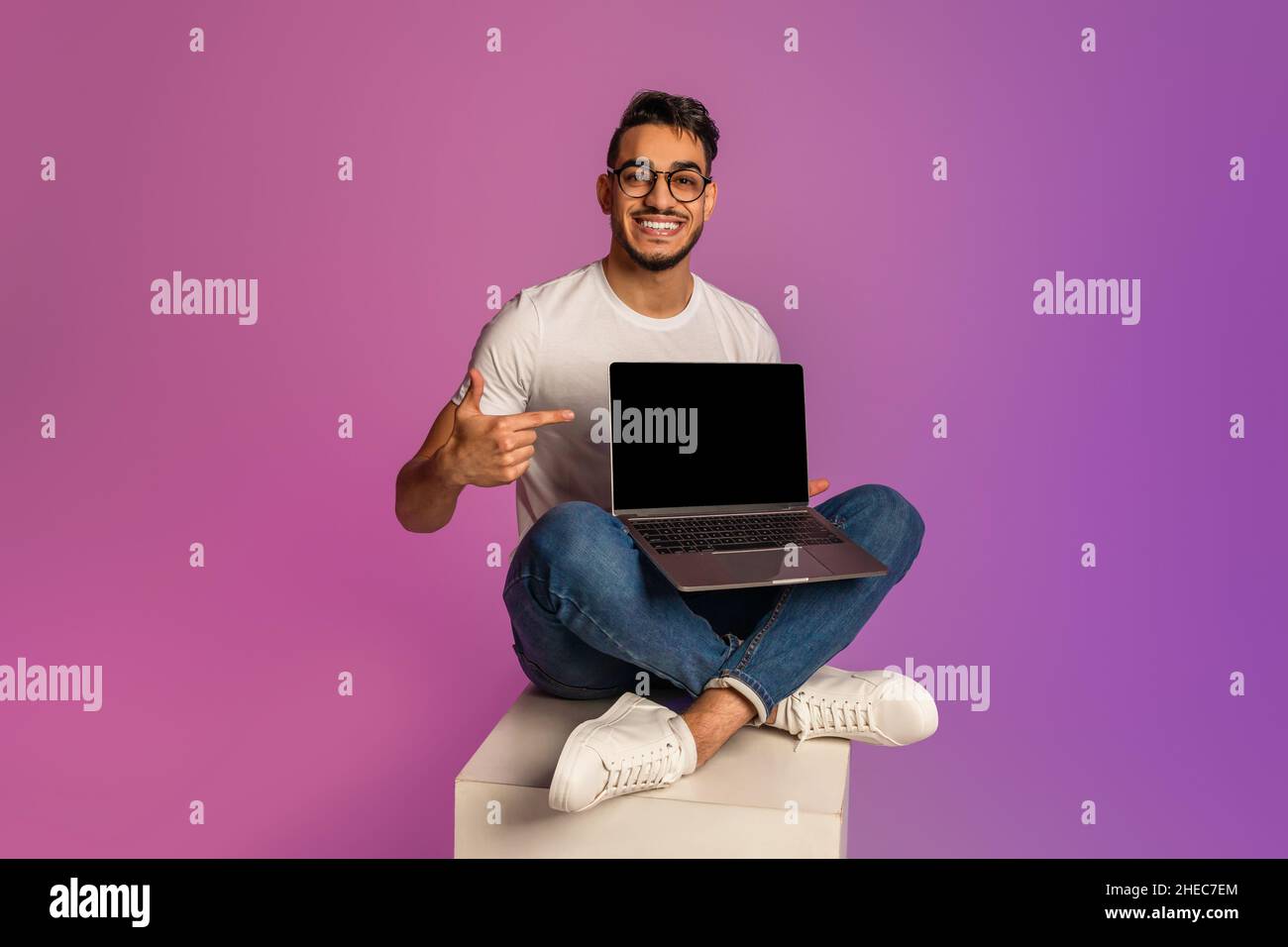 Glücklicher arabischer Mann, der mit einem Laptop überkreuzt sitzt, auf einen leeren Bildschirm zeigt und Raum für Nachahmung in Neonlicht zeigt Stockfoto