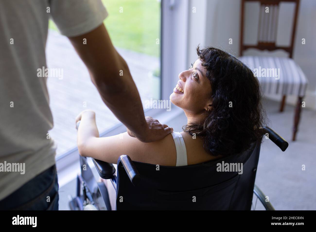 Mittlerer Abschnitt eines Mannes, der zu Hause seine Hand auf die Schulter seiner behinderten Frau im Rollstuhl legt Stockfoto