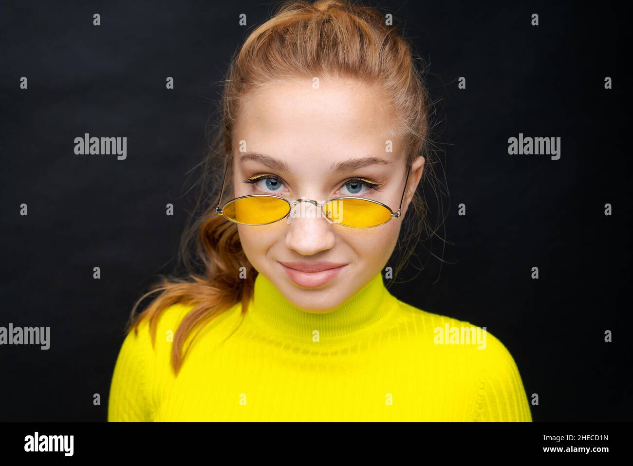 Close-up Portrait glückliches junges Mädchen mit blonden Haaren, schaut durch gelbe Brille, in einem gelben Pullover, ist in guter Stimmung auf schwarzem Hintergrund. Kaukasische Frau Teenager Stockfoto