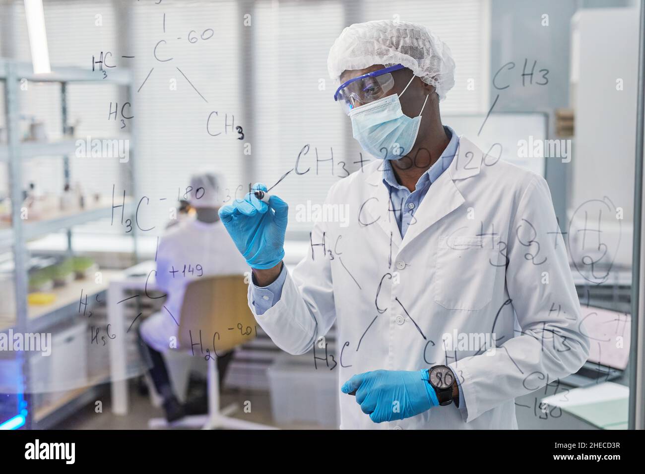 Afrikanischer Chemiker in Schutzkleidung, der chemische Formeln auf Glasplatten schreibt und im Hintergrund mit seinen Kollegen neue Viren untersucht Stockfoto