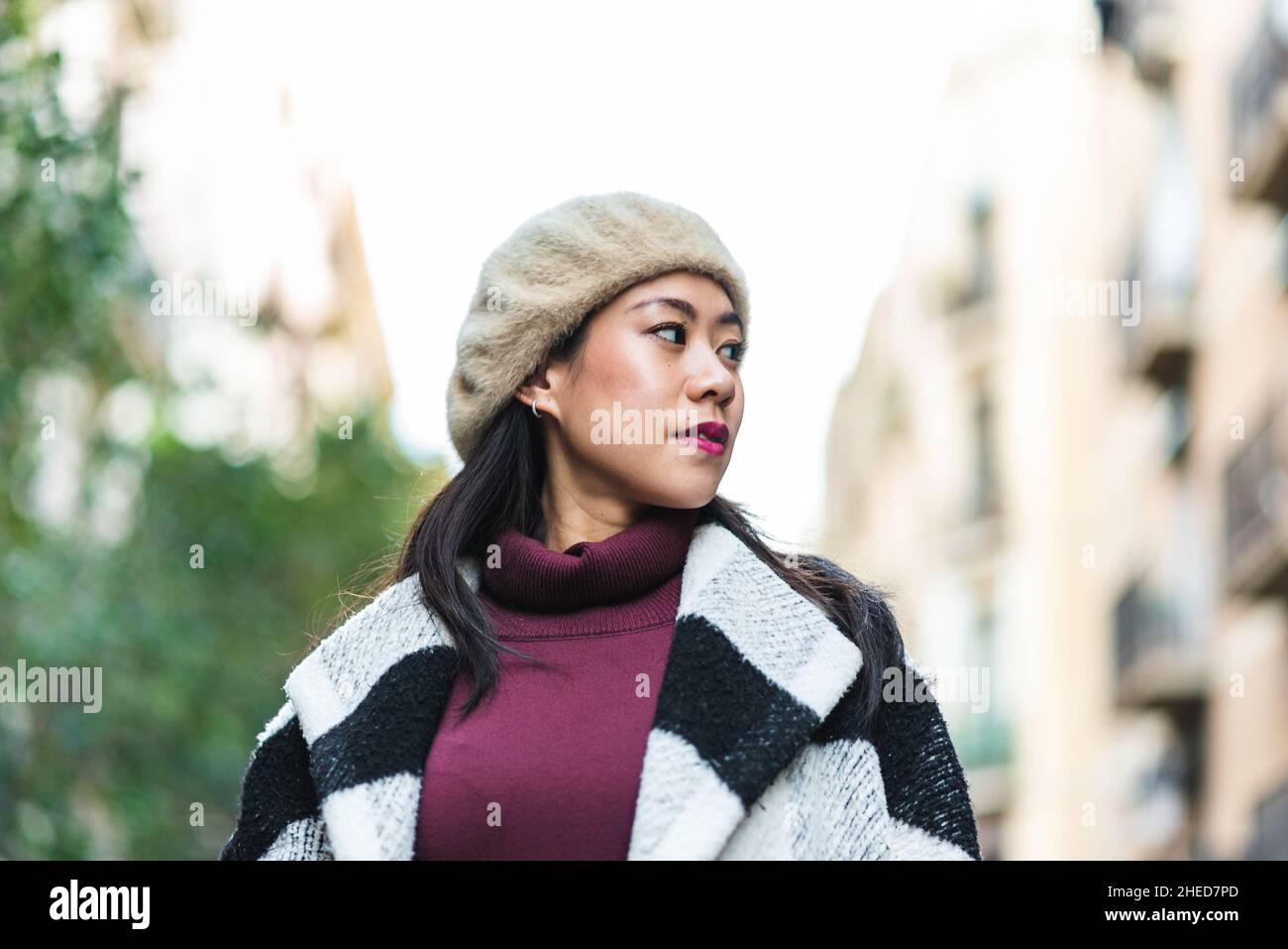 Attraktive chinesische Frau mit schwarzem Haar in Baskenmütze und stylischer warmer Kleidung, die auf der Straße mit Gebäuden vor verschwommenem Hintergrund steht Stockfoto