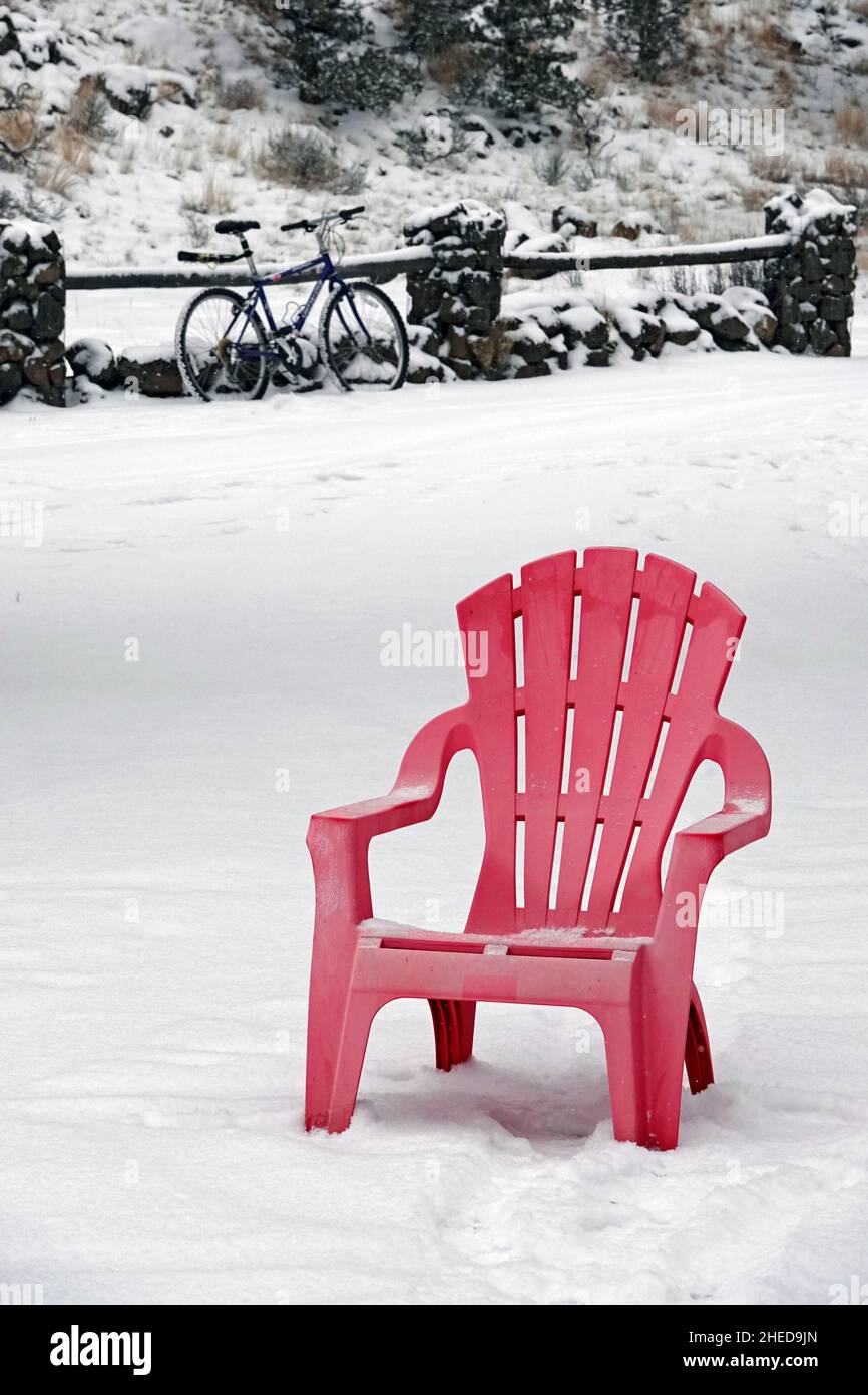Ein roter Rasenstuhl, der während eines Schneesturms im Hof ausgelassen wurde, mit einem uralten Mountainbike, das im Hintergrund an einen Holzzaun gelehnt ist. Stockfoto