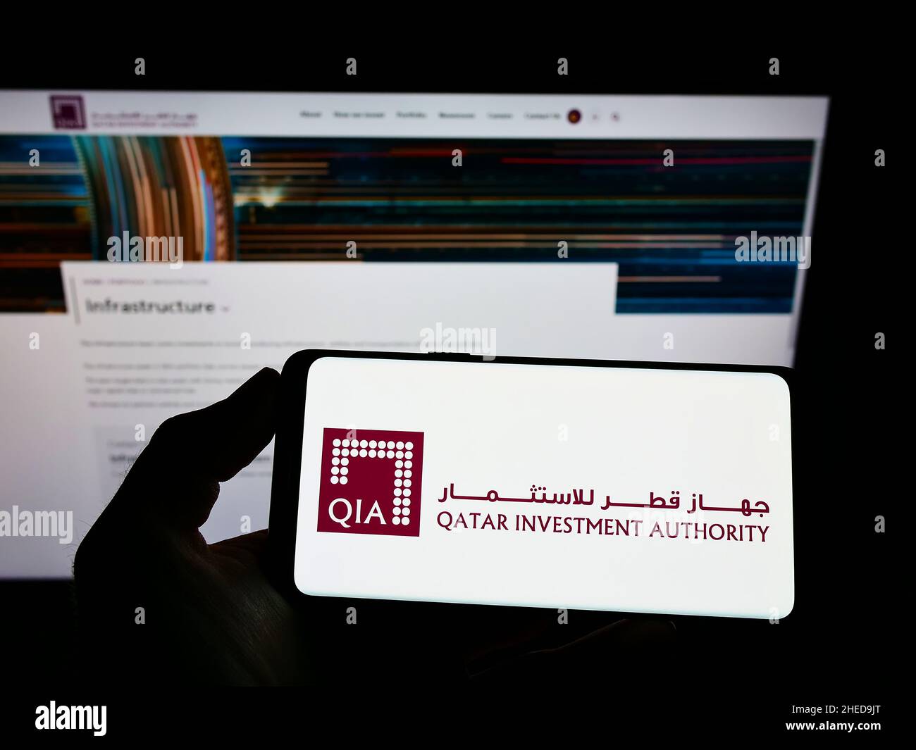 Person, die Mobiltelefon mit Logo des Vermögensfonds Qatar Investment Authority (QIA) auf dem Bildschirm vor der Business-Webseite hält. Konzentrieren Sie sich auf die Telefonanzeige. Stockfoto