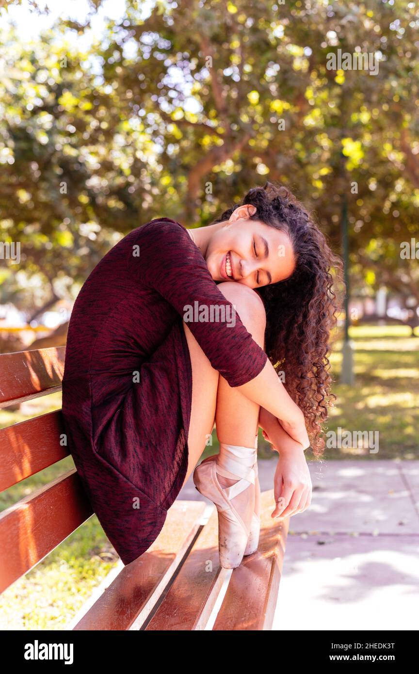 Junge schöne Ballerina in schwarzem Kleid sitzt auf einer Bank in einem Park mit geschlossenen Augen und lächelnd Stockfoto