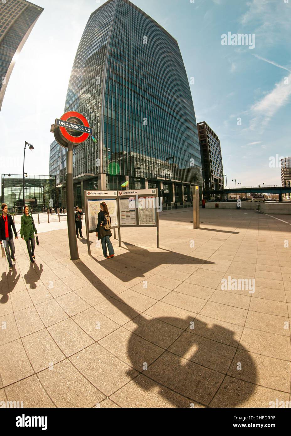 London, Großbritannien - 06. April 2007: Sonne scheint auf unterirdisches Schild am Bahnhof Canary Wharf, vorbeigehende Menschen, hohe Wolkenkratzer im Hintergrund - w Stockfoto