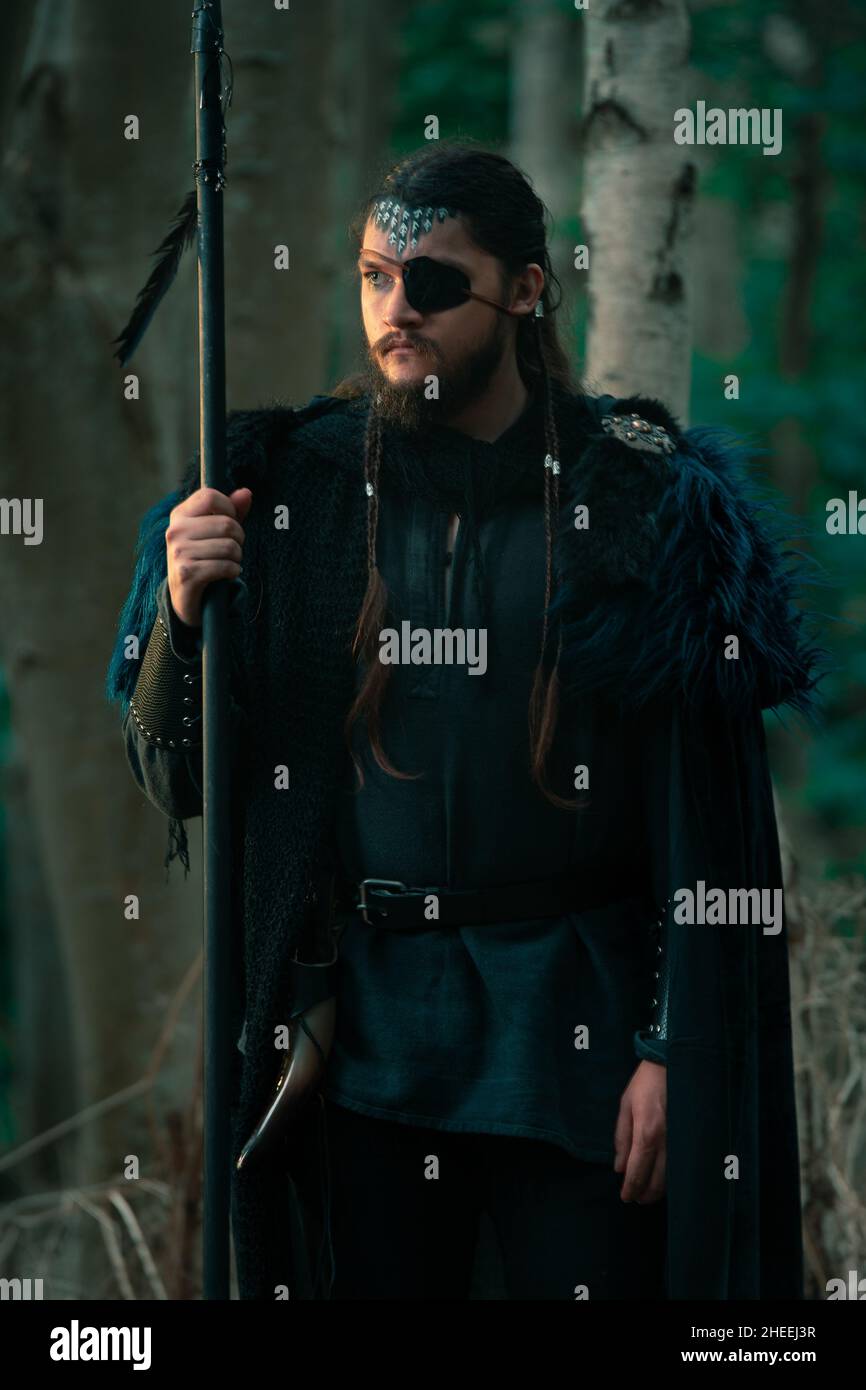 Ernsthafter bärtiger Rüde mit langen schwarzen Haaren im schwarzen wikinger Kostüm mit Augenmuschel, der mit Speer im Wald in der Nähe von Bäumen steht Stockfoto