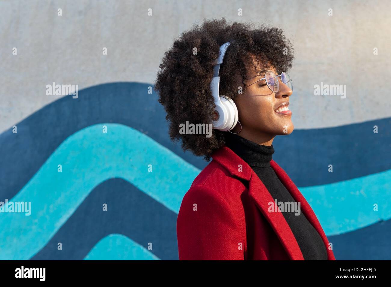 Seitenansicht einer glücklichen afroamerikanischen Frau mit Afro-Frisur und Brillen in schwarzem Rollkragen und roter Jacke, die Podcast kabellos hört Stockfoto