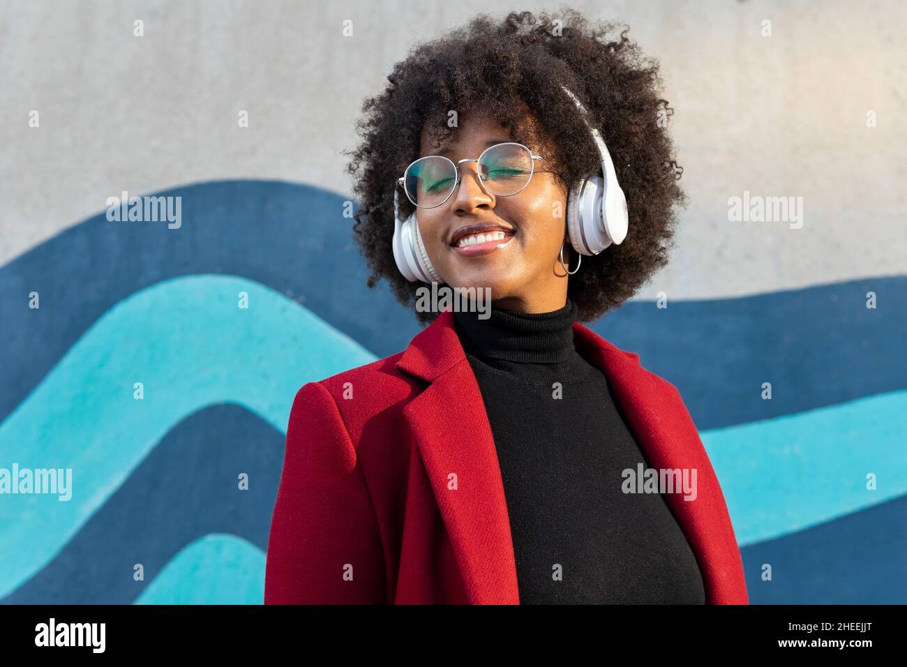 Selbstbewusste, glückliche afroamerikanische Frau mit Afro-Frisur und Brillen in schwarzem Rollkragen und roter Jacke, die Podcast in drahtloser Luftröhre hört Stockfoto