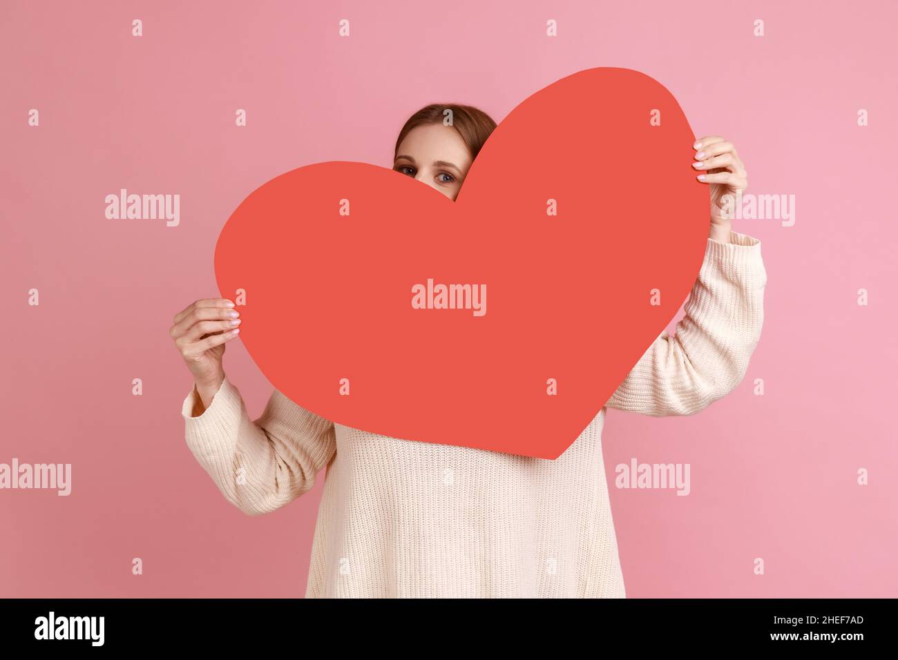 Porträt einer verspielten blonden Frau, die aus dem großen roten Herzen guckt und positive Emotionen ausdrückt, sich verliebt und weißen Pullover trägt. Innenaufnahme des Studios isoliert auf rosa Hintergrund. Stockfoto