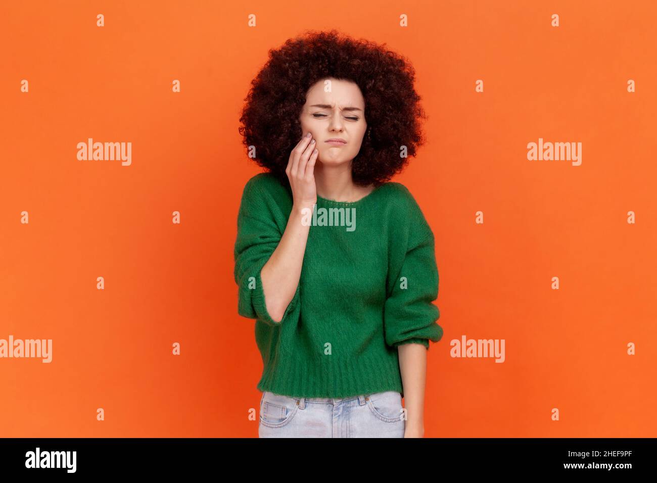 Kranke Frau mit Afro-Frisur trägt einen grünen Pullover im Casual-Stil, der ihre Wange berührt, unter Zahnschmerzen leidet und das Gesicht vor Schmerzen runzelt. Innenaufnahme des Studios isoliert auf orangefarbenem Hintergrund. Stockfoto