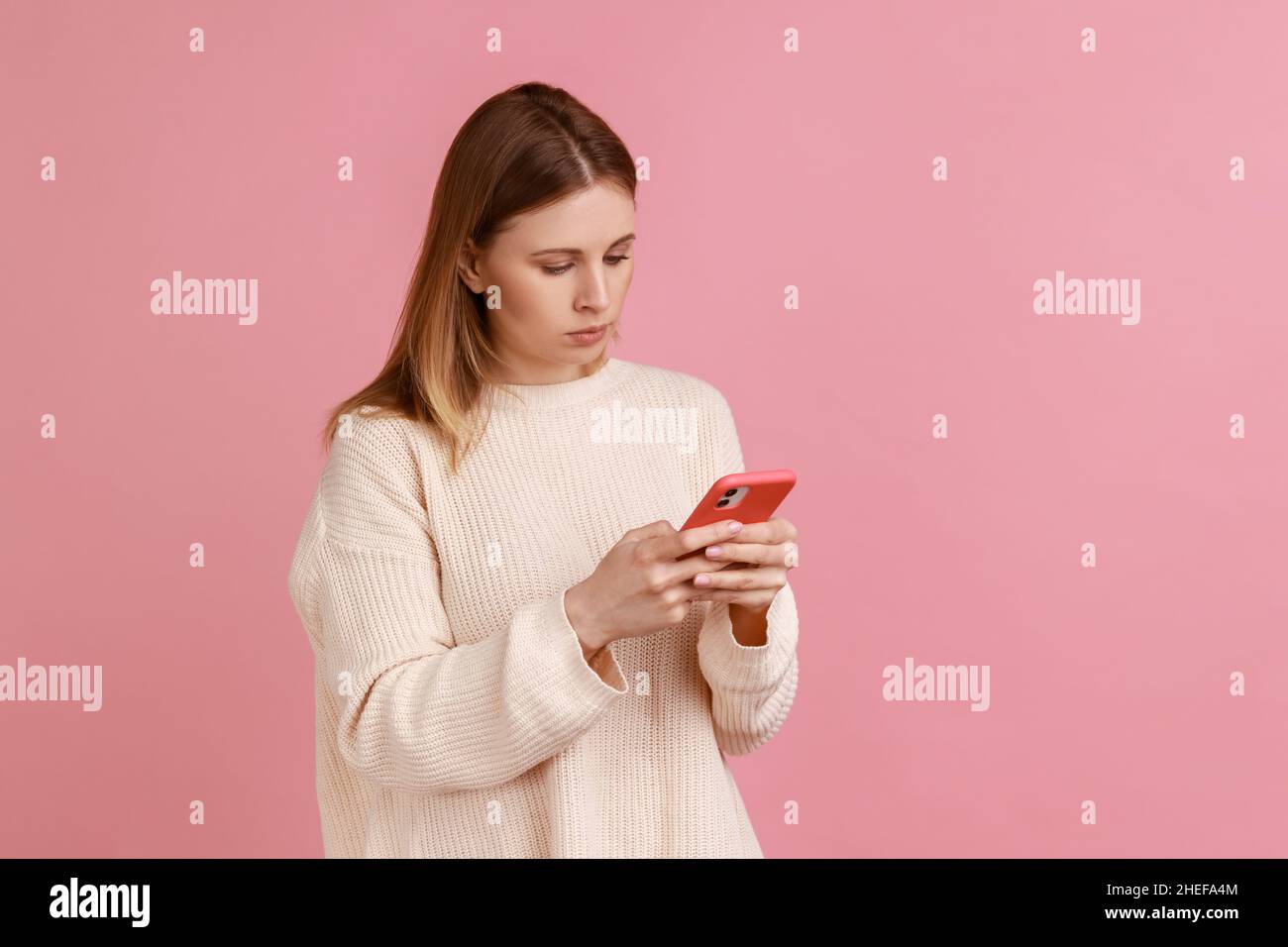 Porträt einer ernsthaft konzentrierten attraktiven blonden Frau, die Mobiltelefon benutzt, auf den Bildschirm des Geräts schaut, eine Nachricht tippt, einen weißen Pullover trägt. Innenaufnahme des Studios isoliert auf rosa Hintergrund. Stockfoto