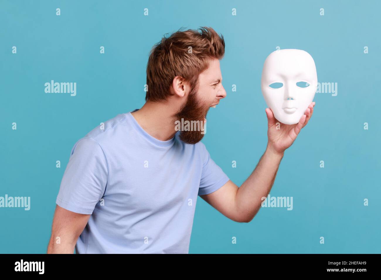 Porträt eines bärtigen Mannes, der wütend schreit, weiße Maske in den Händen hält, seine verborgene Persönlichkeit hasst und negativen Hass ausdrückt. Innenaufnahme des Studios isoliert auf blauem Hintergrund. Stockfoto