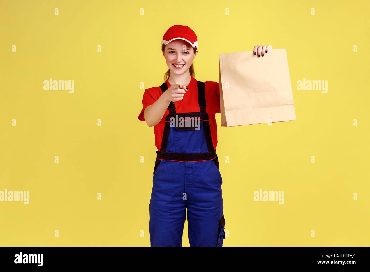 Porträt einer Kurierin, die das bestellte Paket hält, Lebensmittel in einer Papiertüte liefert, den Finger auf die Kamera zeigt, Overalls und Mütze trägt. Innenaufnahme des Studios isoliert auf gelbem Hintergrund. Stockfoto