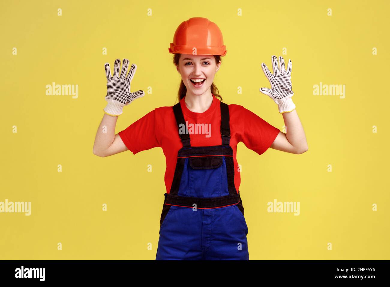 Porträt einer aufgeregten Arbeiterin, die mit erhobenen Armen steht und ihre Handschuhe zum Schutz der Hände zeigt, während die Arbeit Overalls und Schutzhelm trägt. Innenaufnahme des Studios isoliert auf gelbem Hintergrund Stockfoto