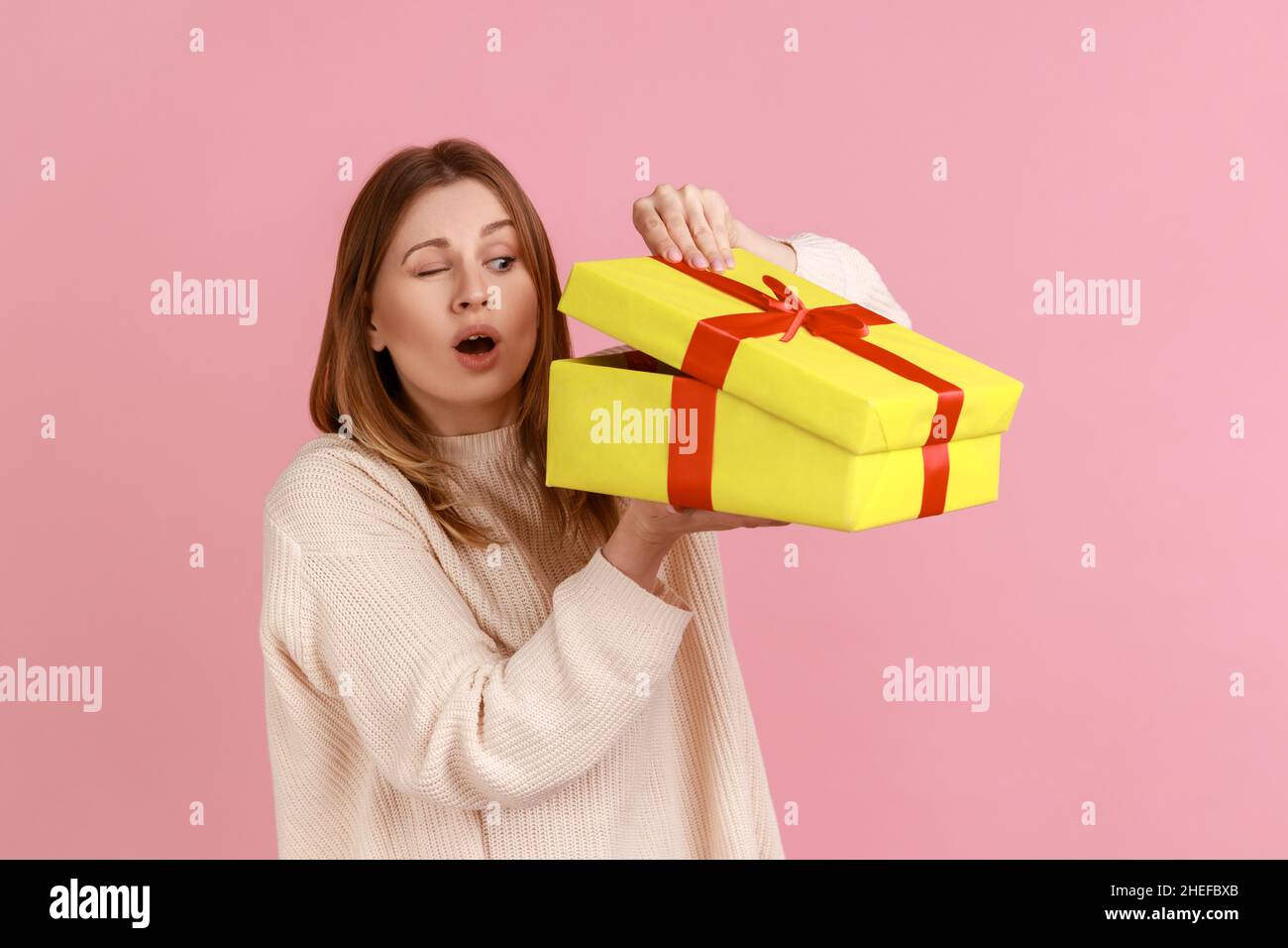 Porträt der neugierigen attraktiven jungen blonden Frau, die in gelben Geschenk-Box, interessiert, was im Inneren, trägt weißen Pullover. Innenaufnahme des Studios isoliert auf rosa Hintergrund. Stockfoto