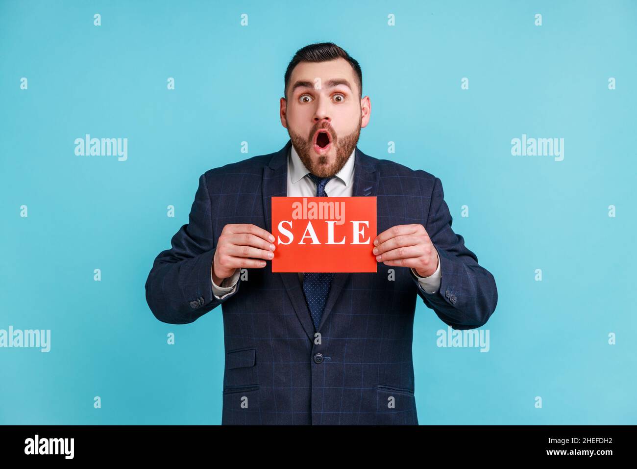 Wow, unglaublicher Einkaufsrabatt. Mann mit Bart trägt offiziellen Stil Anzug zeigt Verkauf Inschrift und mit Staunen. Innenaufnahme des Studios isoliert auf blauem Hintergrund. Stockfoto