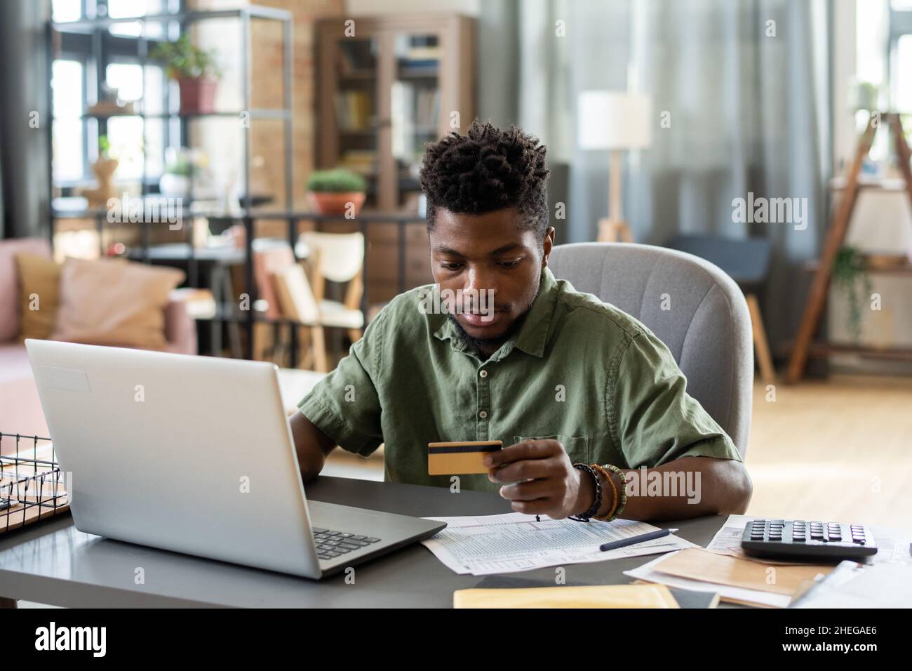 Junger afrikanischer Mann, der die Kreditkarte in der Hand betrachtet, während er vor dem Laptop am Tisch sitzt und für den Online-Kauf bezahlen will Stockfoto