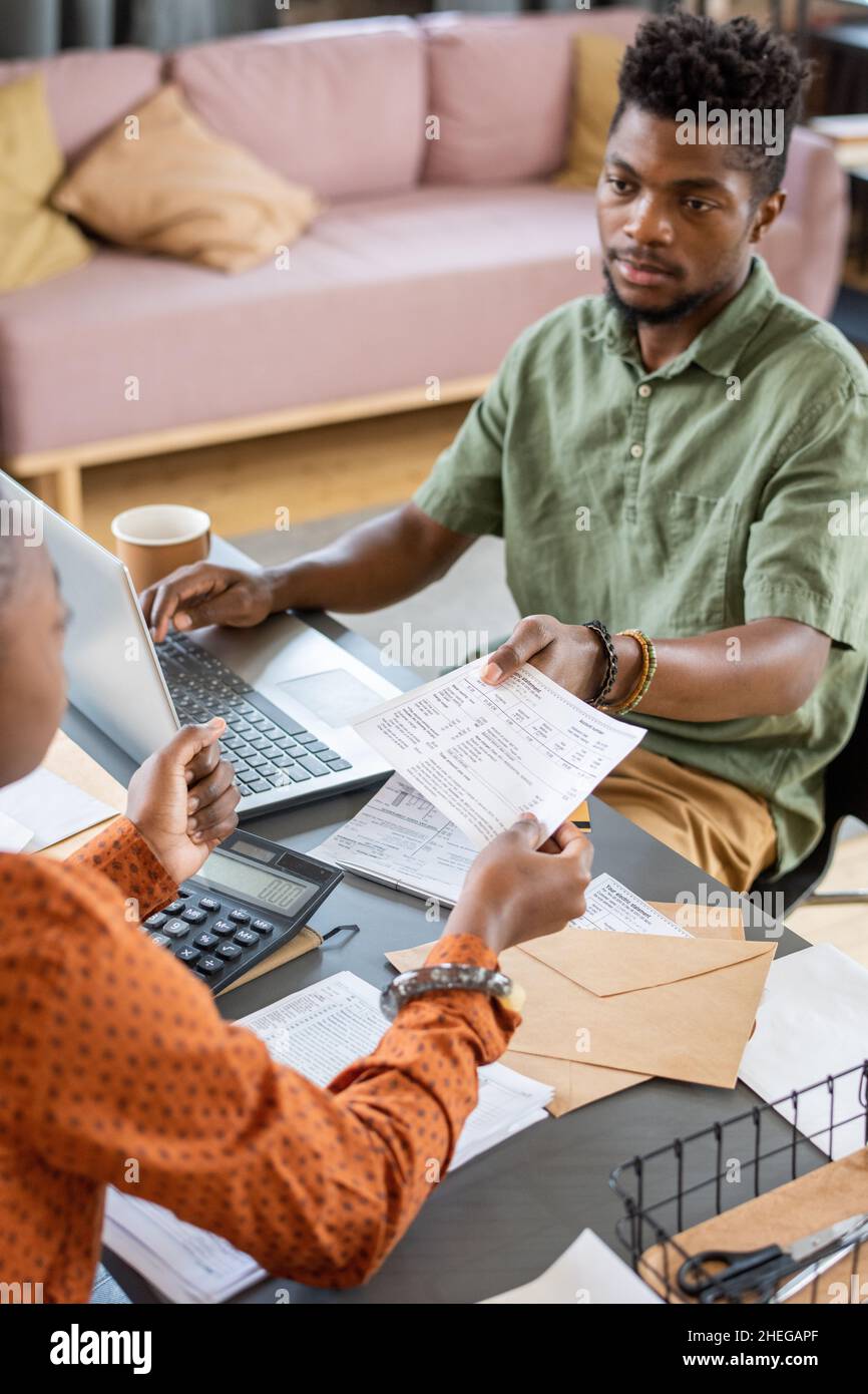 Junger afrikanischer Mann, der Finanzpapier an eine Frau übergibt, während er vor dem Laptop am Tisch sitzt und mit Daten arbeitet Stockfoto