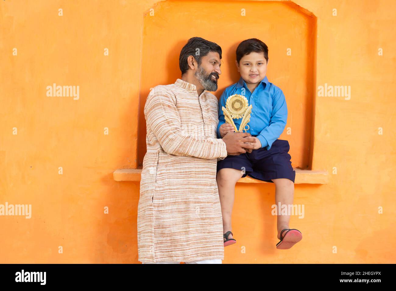 Portrait Junge glückliche indische Vater mit seinem niedlichen Sohn mit Siegerpreis, lächelnder Mann in traditionellem Outfit, der mit dem Jungen in der Schulkleidung Showin steht Stockfoto