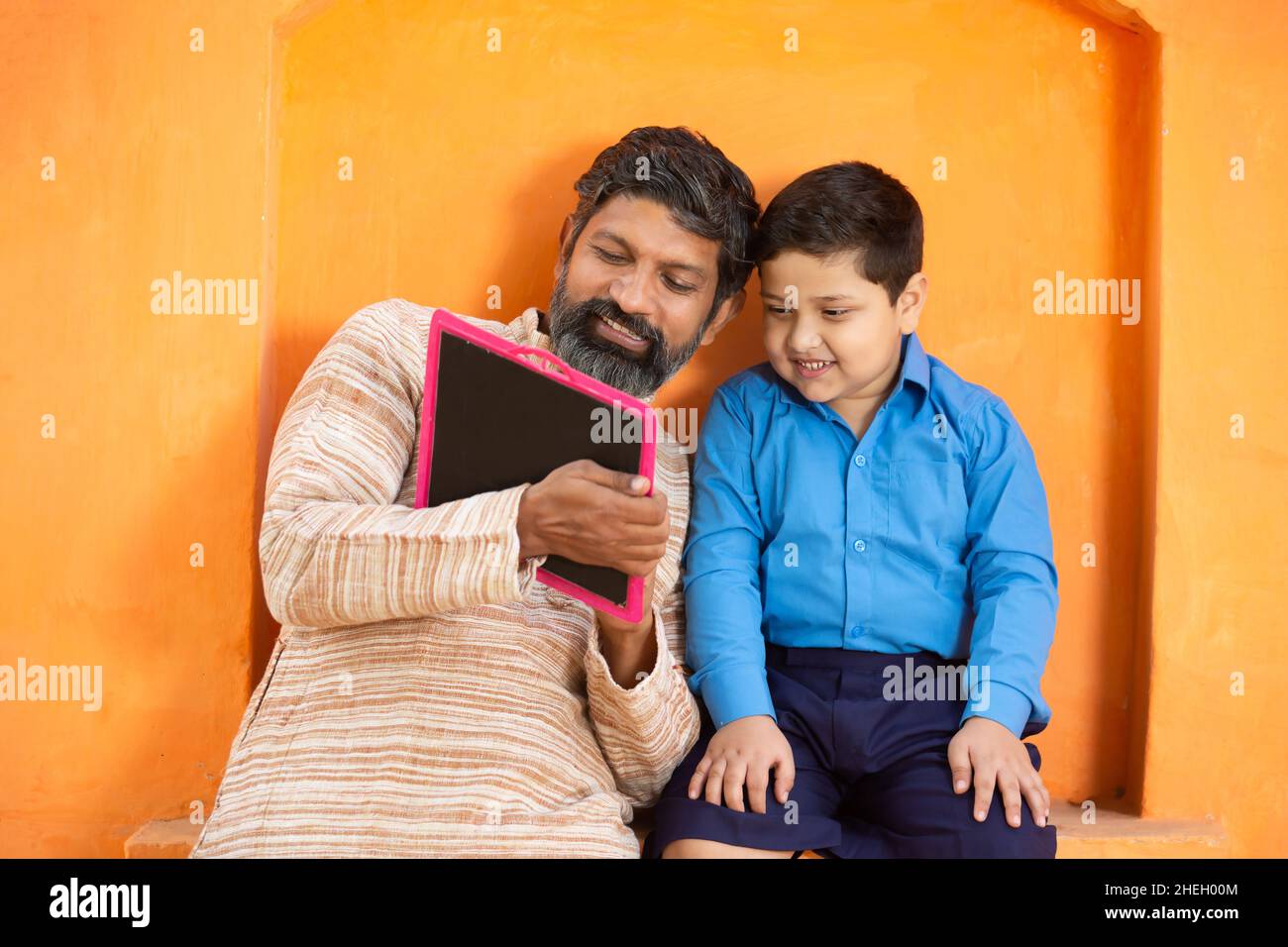 Fröhlicher indischer Vater und sein süßer kleiner Sohn in Schuluniform schreiben auf Schiefertafel vor orangefarbenem Hintergrund, entzückendes Grundkind mit Bartmann Stockfoto