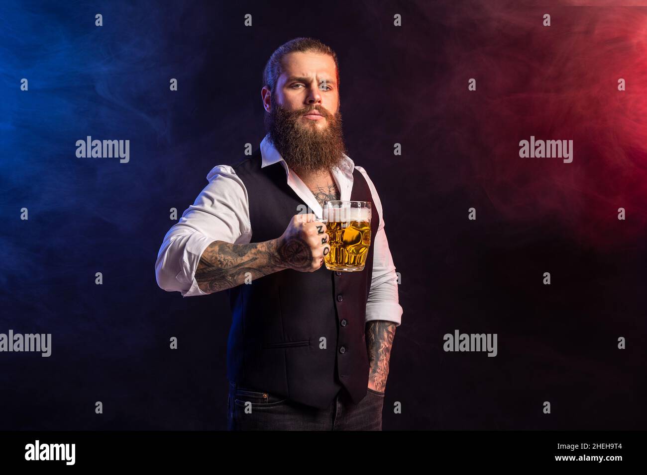 Halb langer bärtiger Mann mit Hipster, der einen Becher Bier in der Tasche hält und über dem Rauchhintergrund auf die Kamera blickt Stockfoto