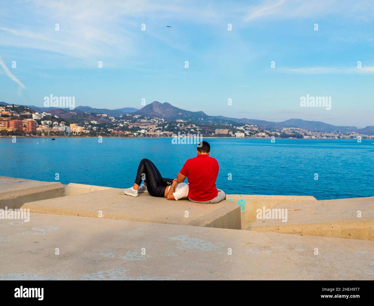 Junges Paar, das auf dem Rücken auf einem Dock im Hafen sitzt und die Küste der Bucht von Malaga betrachtet. Stockfoto