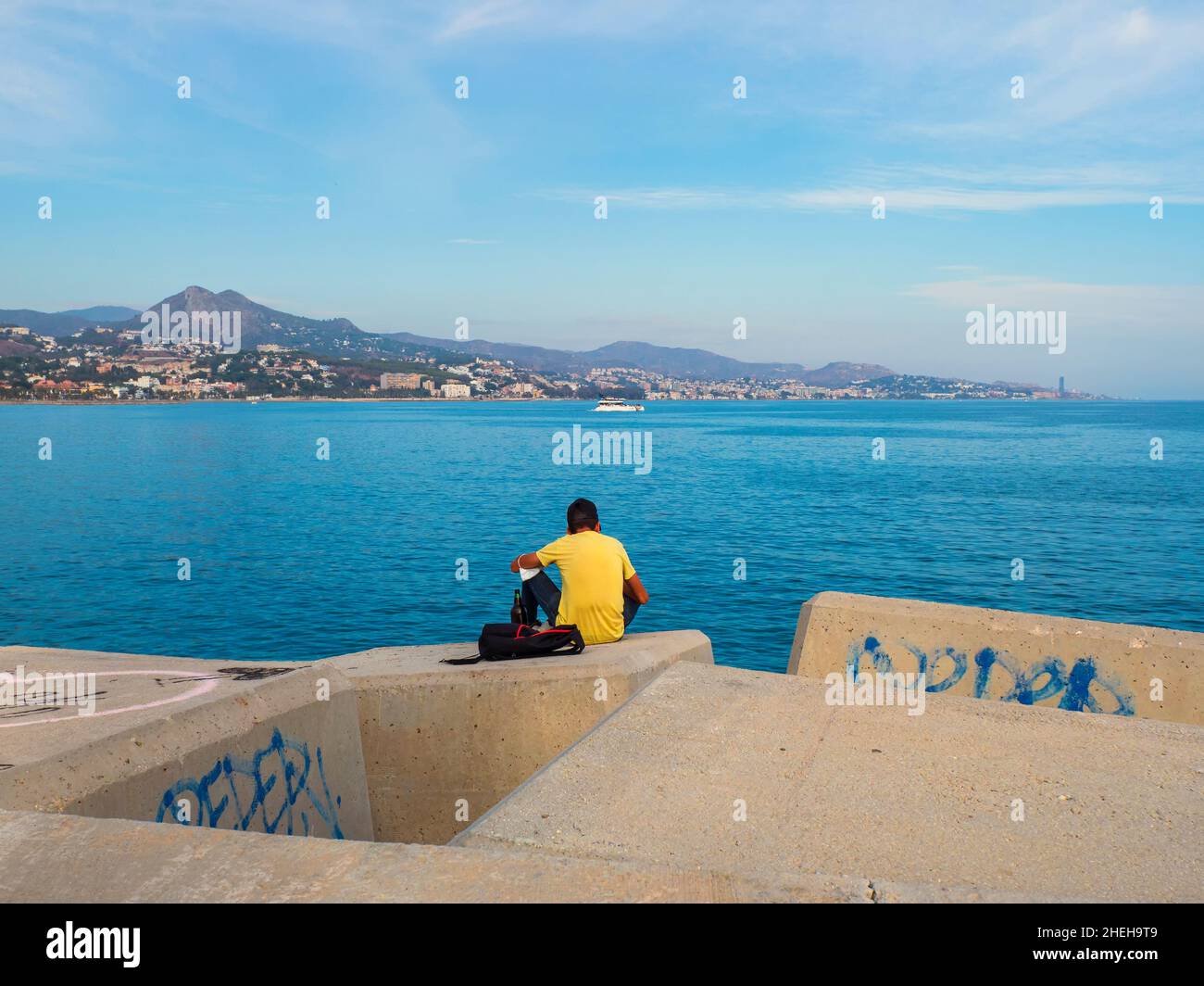 Junger Mann sitzt auf dem Rücken auf einem Deich im Hafen und blickt auf die Küste der Bucht von Malaga. Stockfoto