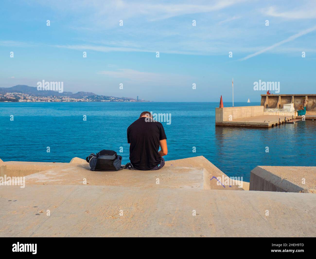 Junger Mann sitzt auf dem Rücken auf einem Deich im Hafen und blickt auf die Küste der Bucht von Malaga. Stockfoto