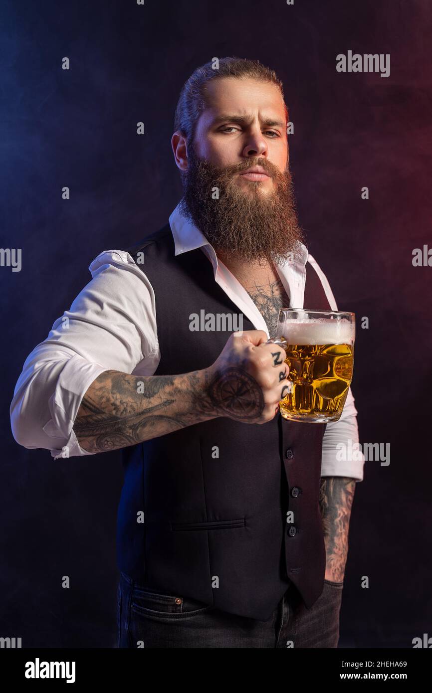 Porträt eines Bartmenschen, der einen Becher Bier in seinen Tätowierungen hält und die Kamera auf schwarzem Hintergrund anschaut Stockfoto
