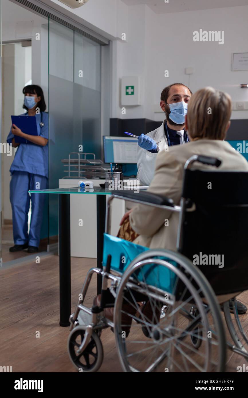 Arzt trifft sich mit einem alten Patienten im Rollstuhl bei der Untersuchung während einer covid 19 Pandemie. Spezialist, der Rentner mit körperlicher Behinderung in der Arztpraxis unterstützt und berät. Stockfoto