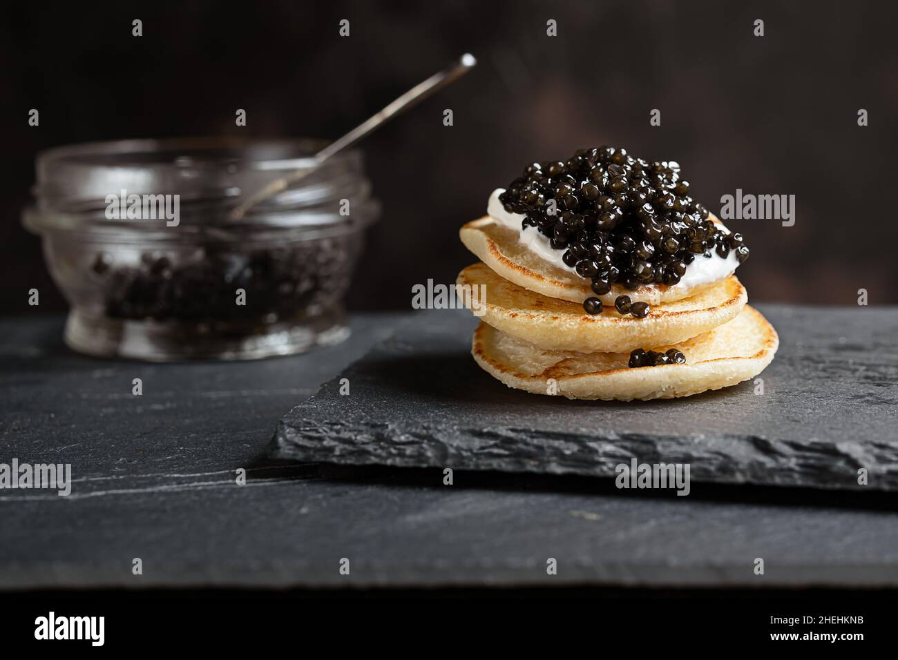 Schwarzer Stör-Kaviar auf kleinen Pfannkuchen blinis mit saurer Creme und einem Glas mit Kaviar auf einem Schiefer Stockfoto