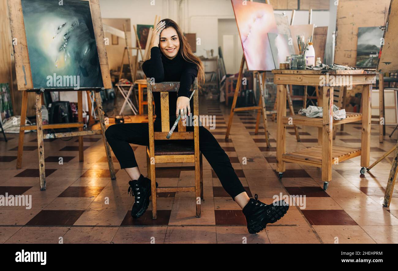Fröhliche Künstlerin lächelt fröhlich auf die Kamera, während sie auf einem Stuhl in ihrer Werkstatt sitzt. Selbstständige Malerin mit einem Pinsel Stockfoto