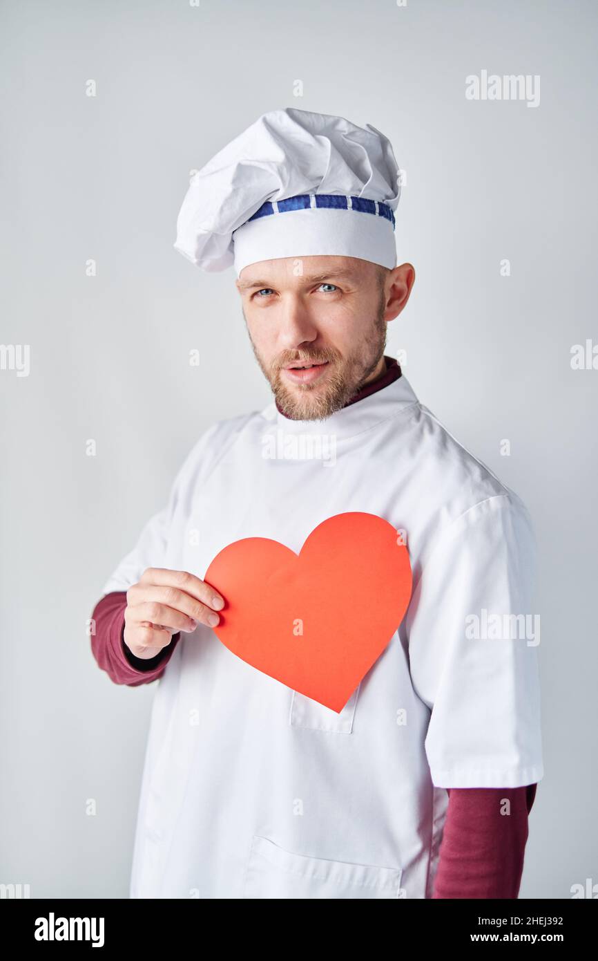 Bärtiger Chefkoch oder Bäckermann in weißer Uniform mit rotem Papierherz als St. Valentinstag-Konzept. Koch hält ein rotes Herz vor der Arbeitskleidung. Hochwertige Fotos Stockfoto