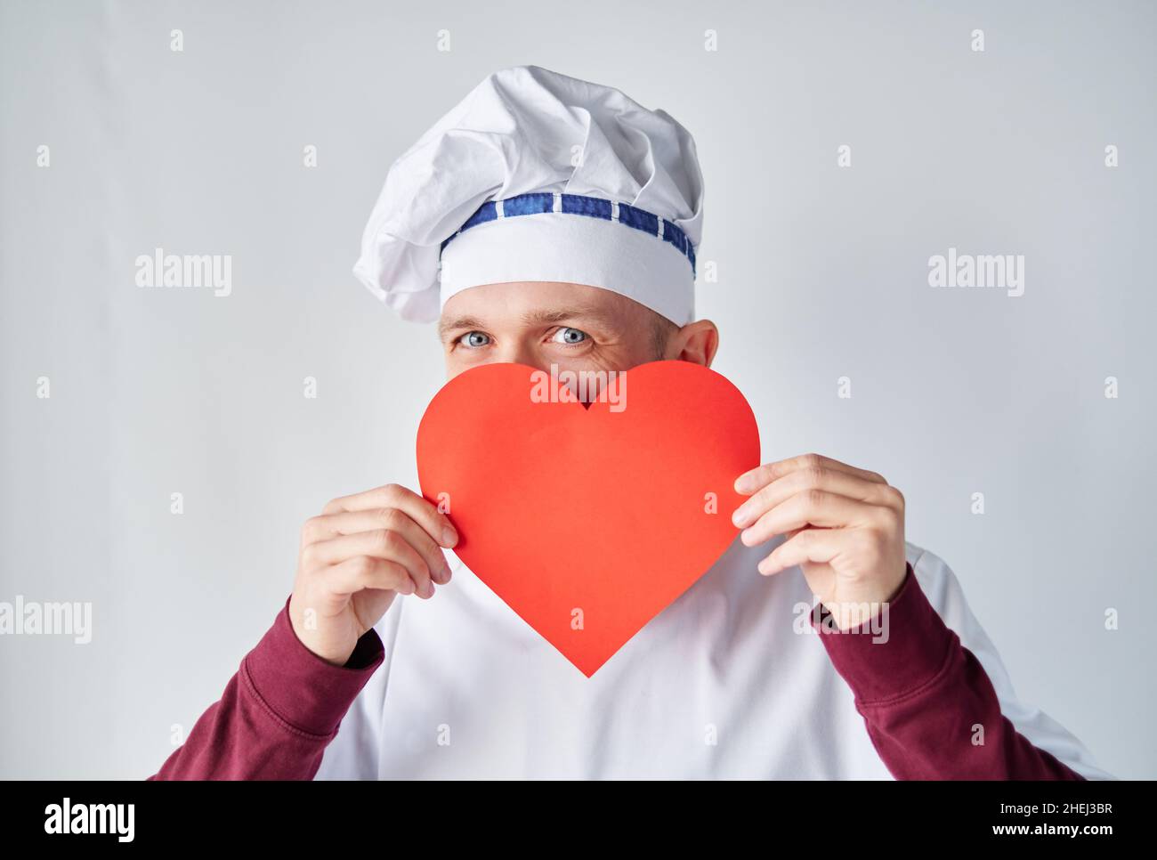 Valentinstag: Attraktive bärtige Koch Koch oder Bäcker Mann in weißer Uniform mit rotem Papier Herz als. Koch hält ein rotes Herz vor der Arbeitskleidung. Liebe oder Beziehungskonzept Stockfoto