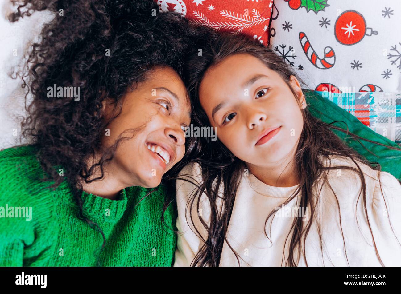 Afroamerikanische Mutter mit lockigen Haaren umarmt die brünette Tochter und lacht, während sie an Weihnachten gemütlich im Wohnzimmer auf Kissen liegt Stockfoto