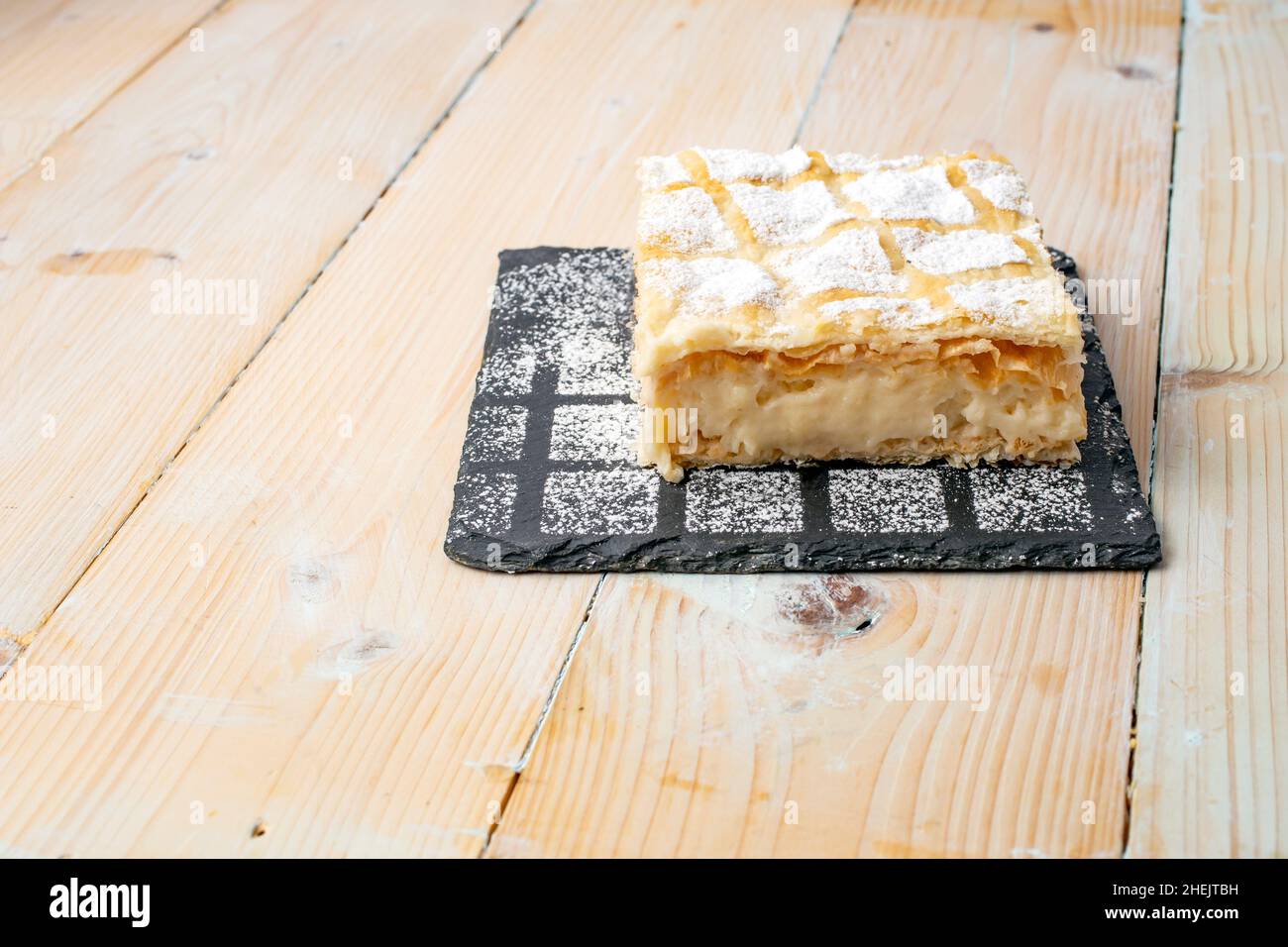 Cremeschnit oder Cremsnit Kuchen auf einer quadratischen schwarzen Steinfliesen Textur mit bestreuten Zuckerpulver und einem weißen gewaschenen Holzbretttisch Stockfoto