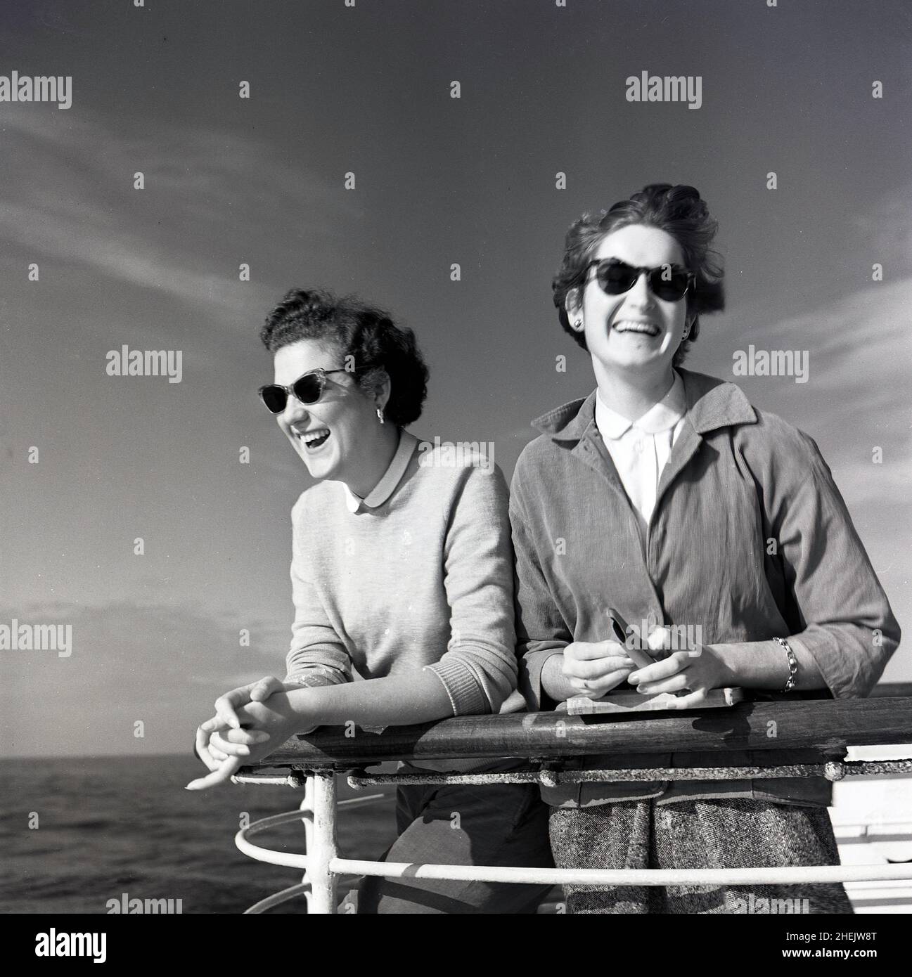 1950s, historisch, auf dem Meer, zwei junge Damen genießen die Brise, die an einem Geländer auf dem Deck eines Union Castle-Kreuzfahrtschiffs steht. Stockfoto