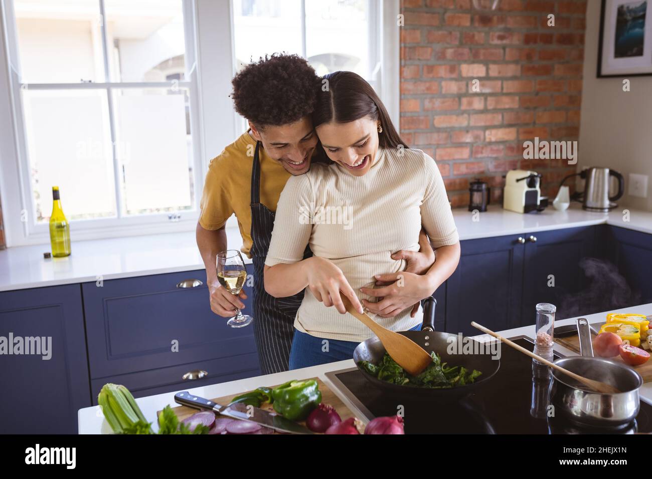 Glücklicher junger Mann hält Getränk umarmt Freundin von hinten Kochen Mahlzeit in der Küche zu Hause Stockfoto