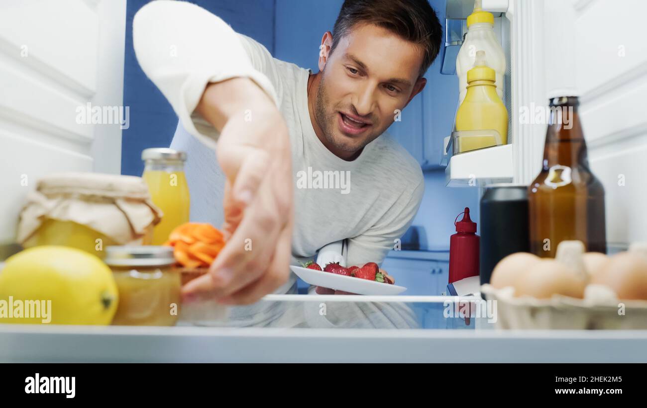 Fröhlicher Mann, der leckeren Cupcake und Erdbeeren aus dem Kühlschrank mit Getränken, Eiern und Behältern mit Essen nahm Stockfoto