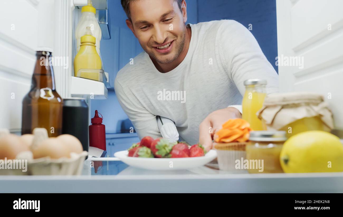 Fröhlicher Mann, der reife Erdbeeren aus dem Kühlschrank mit Getränken, Saucen, Eiern und Gläsern zum Essen nimmt Stockfoto