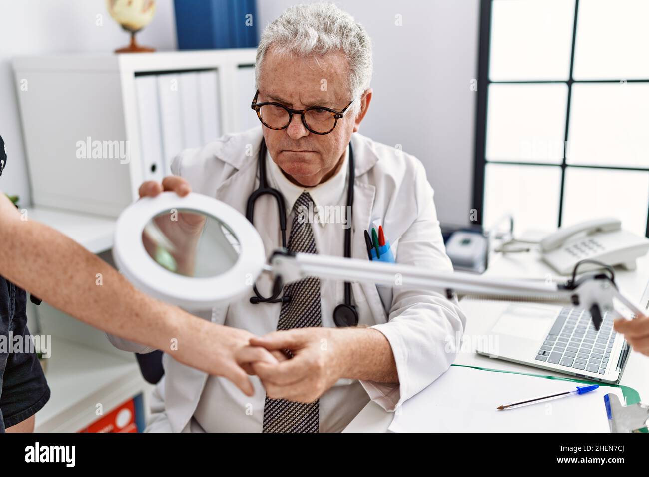 Grauhaariger Mann mittleren Alters, der in dermatologischer Uniform den Hautarm mit einer Lupe in der Dermatologie-Klinik untersucht Stockfoto