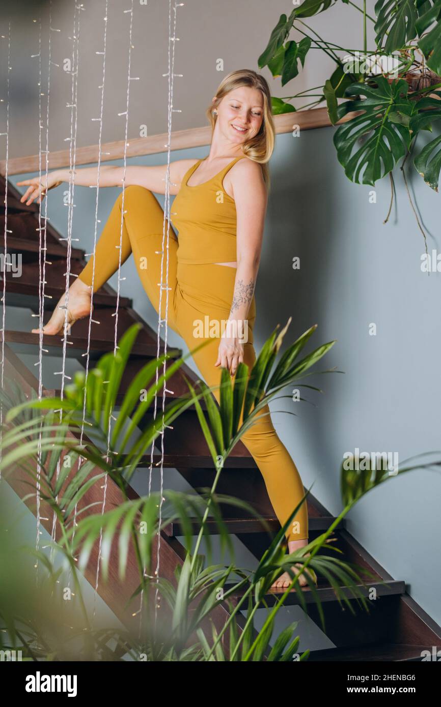 Blonde Frau mit gelbem Oberteil und Leggings macht vertikale Spalten, die sich auf einer Holztreppe in der Nähe von Girlande und grünen Pflanzen aufwärmen Stockfoto