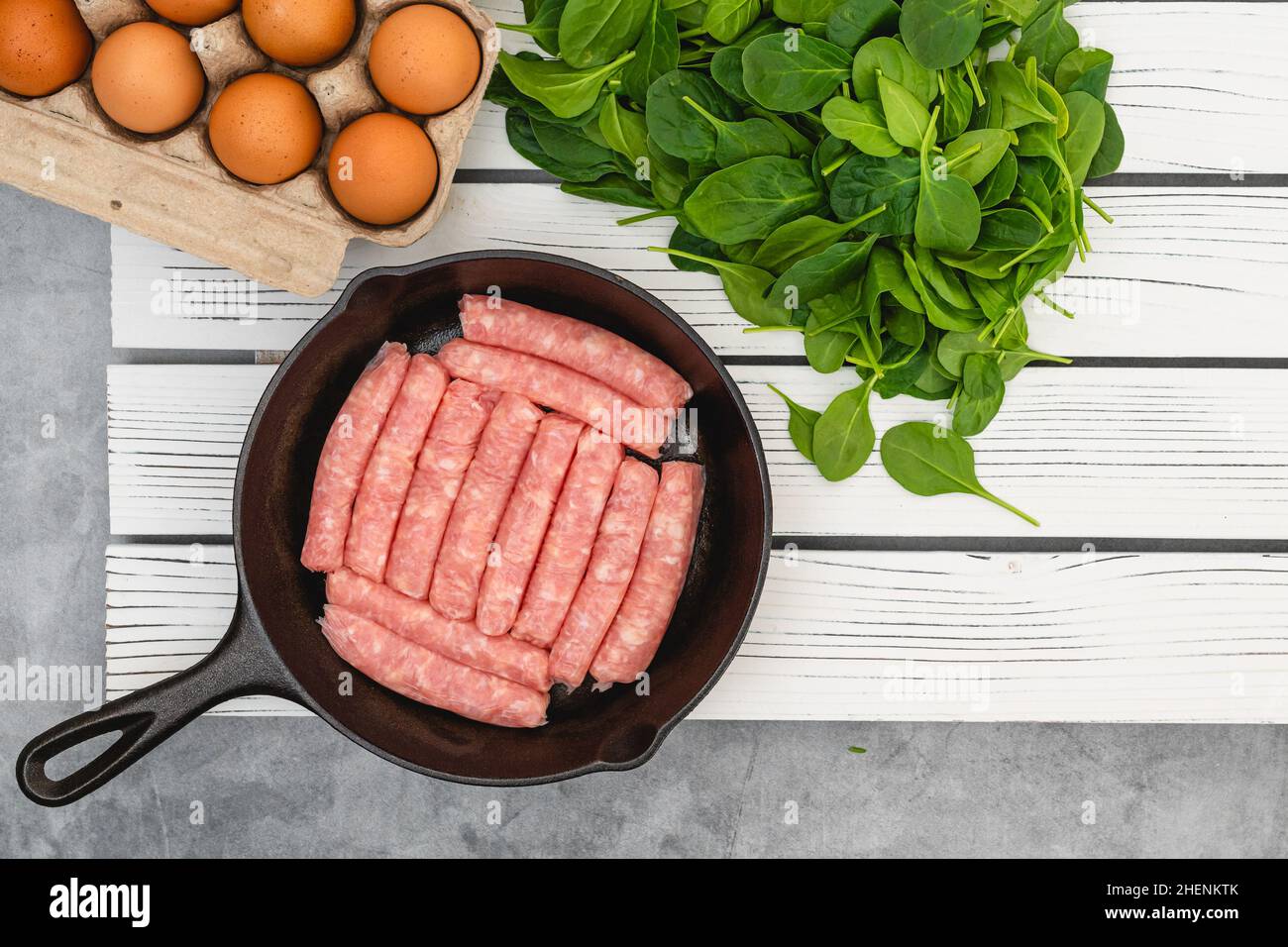 Rohe Hähnchenfrühstückswürste auf der Bratpfanne, Eier und Spinat. Zutaten zum Frühstück aus nächster Nähe auf weißem Holzbrett auf Küchentisch, flach liegend Stockfoto