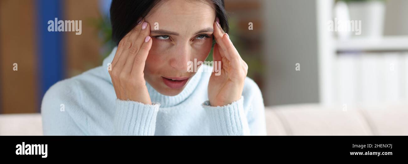 Eine Frau mit Kopfschmerzen hält die Hände an Schläfen, während sie auf der Couch sitzt Stockfoto