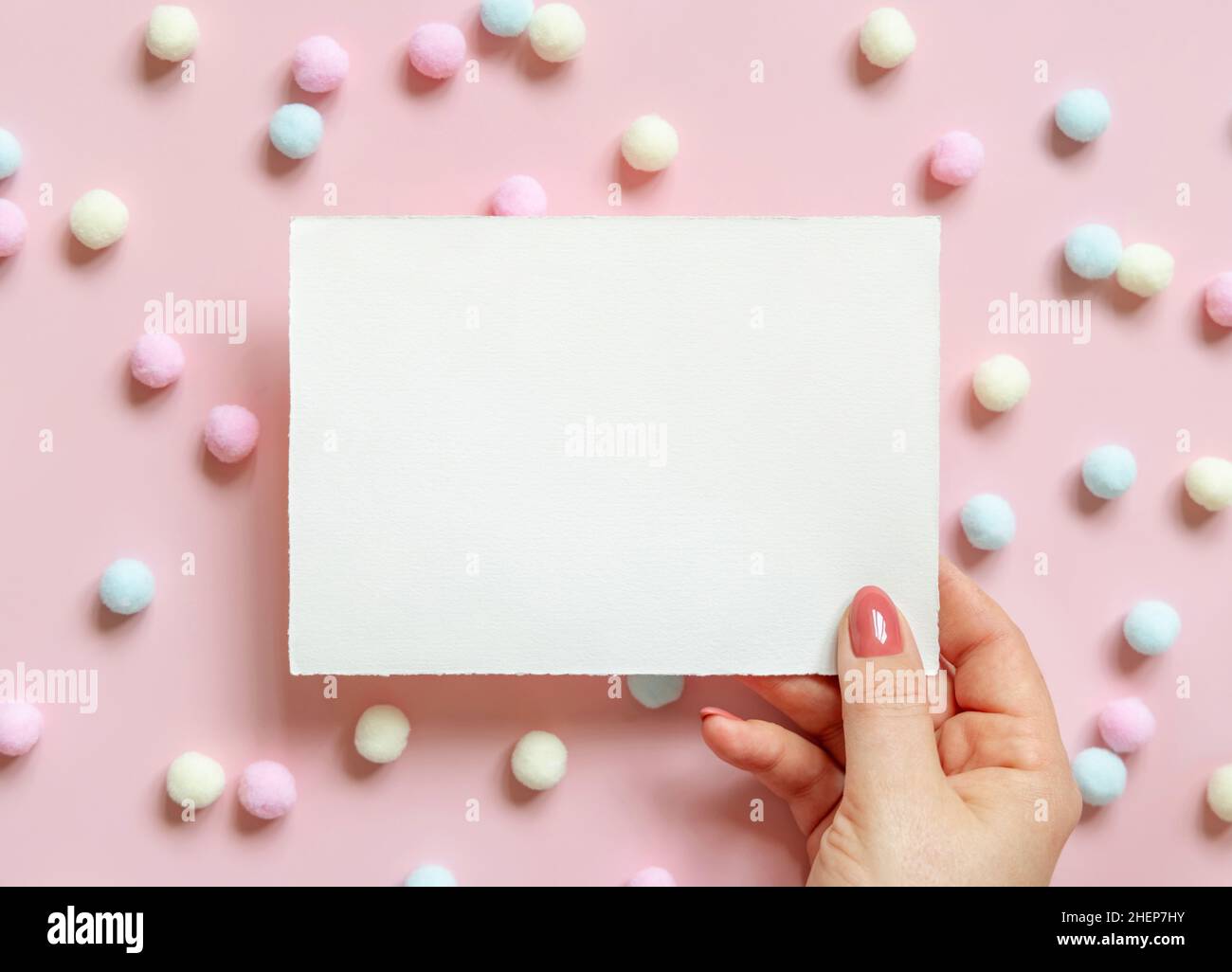 Hand mit leerer Karte in der Nähe von pastellfarbenen Pom-Poms auf rosa Draufsicht. Romantische Szene mit leeren horizontalen Karte mockup flach legen, Platz für Text. Valentines, Sp Stockfoto