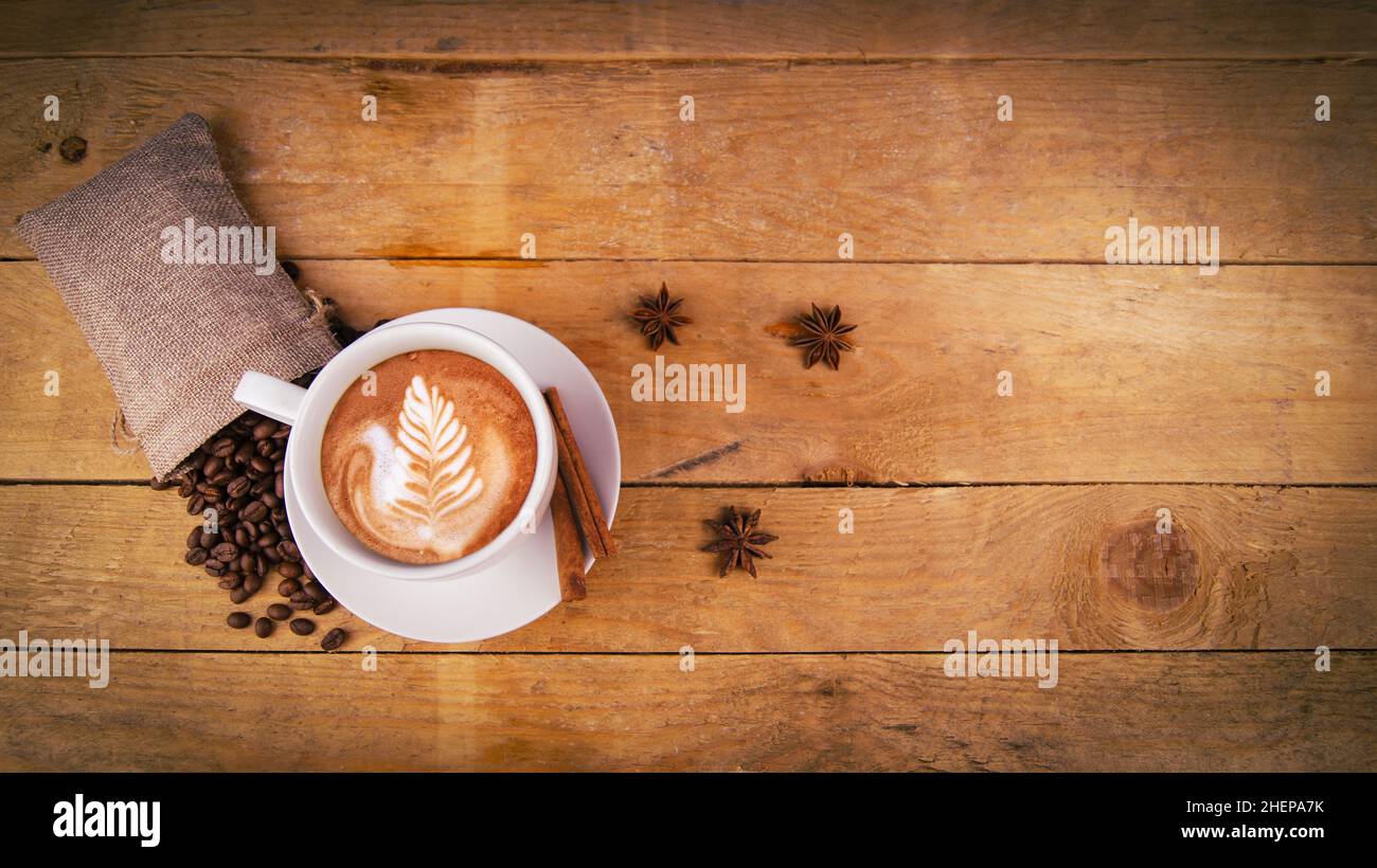 Blick auf die Kaffeetasse von oben auf einem alten Holztisch. Blattmuster auf Cafe Latte. Anissterne, Kaffeebohnen im Beutel und Zimt zur Dekoration. Schön, organisch, Stockfoto