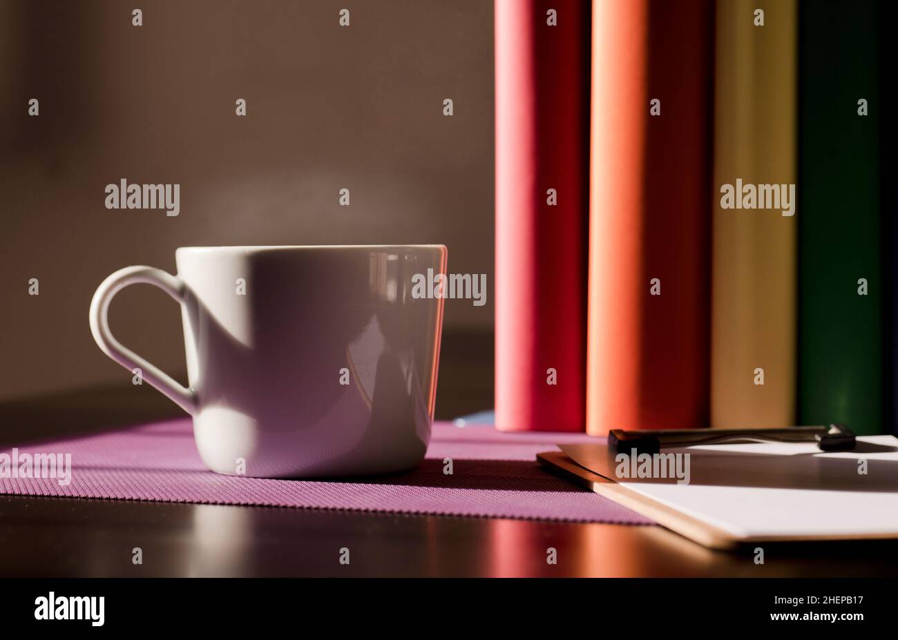 Weißer Keramikbecher mit heißem Kaffee auf purpurfarbenem Schreibtisch und Büchern mit lgbt-Bewegungsfarben Stockfoto