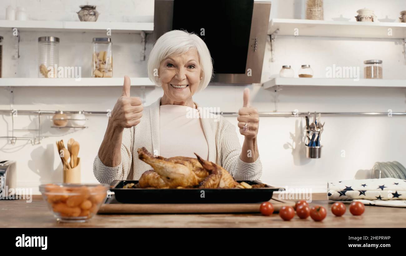 Glückliche Frau, die den Daumen nach oben zeigt, neben gebratenem truthahn und frischem Gemüse auf dem Küchentisch Stockfoto