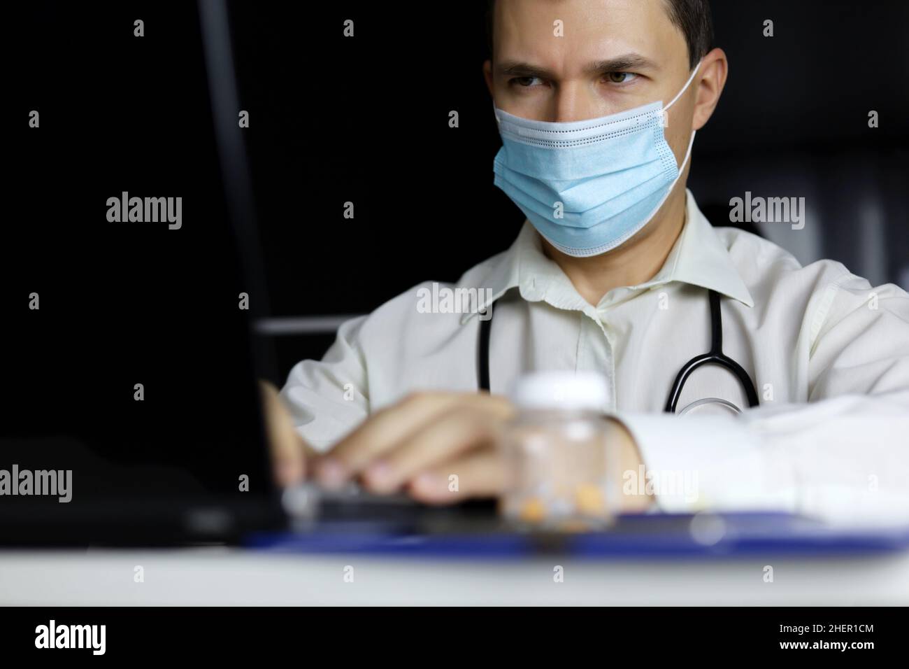 Arzt in Einwegmaske arbeitet am Laptop im Büro. Arzt schreibt Rezept, Konzept der medizinischen Untersuchung, Ausfüllen der Krankengeschichte Stockfoto