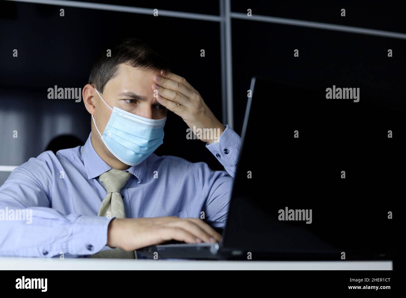 Coronavirus- und Fiebersymptome, Mann in Gesichtsmaske und Bürokleidung legte seine Hand auf die Stirn, sitzend am Laptop. Kranker Büroangestellter Stockfoto